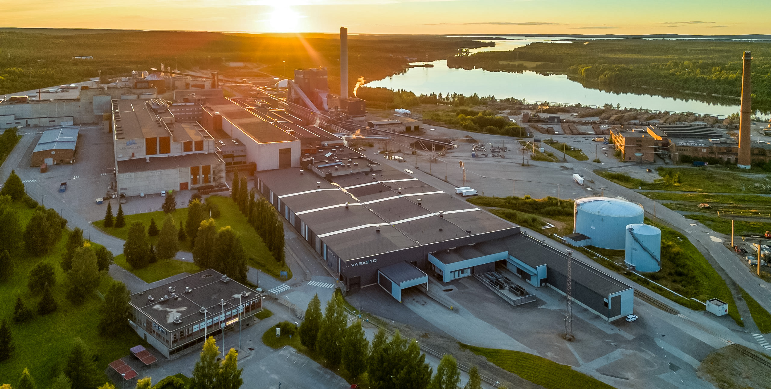Figure 2: Aerial view photograph of the Kajaani data centre. In the foreground the data centre buildings are visible, clearly resembling an old paper mill. In the background a lake and forest are visible. The photo was taken at sunset, giving everything a soft orange glow.